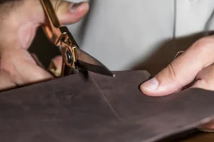 Close-up of a person cutting brown PU leather with gold scissors, showcasing detailed craftsmanship and material preparation used in handbag fabrics and materials production.