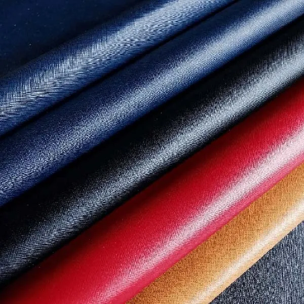 A detailed close-up shot of Saffiano leather, showcasing several folded swatches in rich colors. From top to bottom, the colors are a deep navy blue, a classic black, a vibrant red, and a warm tan. The image highlights the distinctive cross-hatch texture and subtle sheen characteristic of Saffiano leather, emphasizing its luxurious and durable quality.