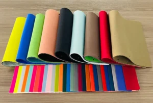 A vibrant collection of colorful PVC fabric swatches laid out on a wooden surface, showcasing material options for bags. The image features large, rolled samples in various colors like blue, red, and black behind a complete swatch card of smaller, vibrant color chips, illustrating the choices available in 'An Ultimate Guide To PVC Fabric For Bags: What You Must Know'.