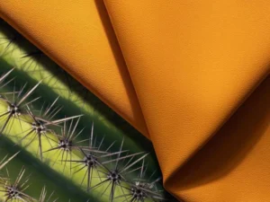 A close-up, high-angle shot shows two folded pieces of soft, orange-yellow cactus leather lying next to the green, spiny arm of a cactus plant. The texture of the vegan leather mimics that of traditional animal leather, highlighting the contrast between the raw, prickly source and the final smooth, luxurious product.