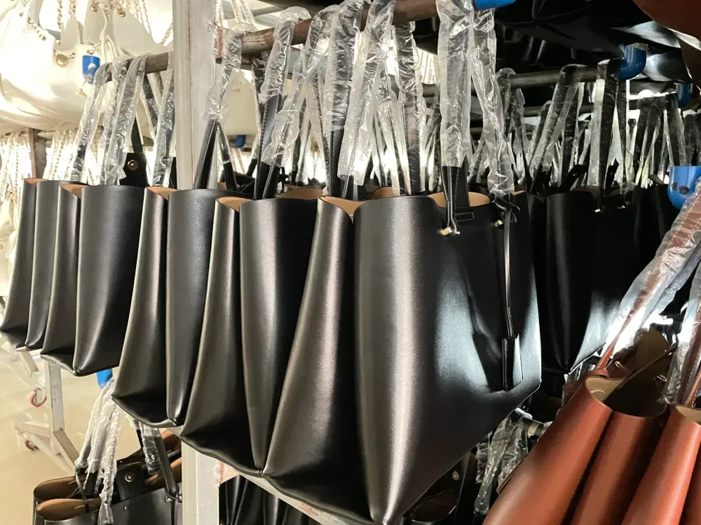 A shot from a factory or warehouse, showing many identical black leather tote bags hanging neatly in rows on a metal rack. The handles of each bag are wrapped in protective clear plastic, indicating they are new products just finished or ready for shipment.