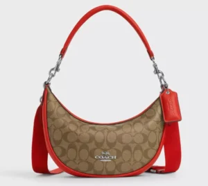 A studio shot of a Coach crescent-shaped hobo bag against a light gray background. The bag features the brand's classic brown signature monogram canvas, accented with vibrant red leather trim. It is shown with two straps: a thin red leather shoulder strap and a thicker, wide red webbed strap. A red leather Coach hangtag and silver hardware complete the design.