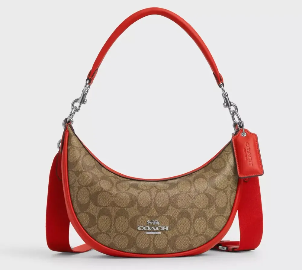 A studio shot of a Coach crescent-shaped hobo bag against a light gray background. The bag features the brand's classic brown signature monogram canvas, accented with vibrant red leather trim. It is shown with two straps: a thin red leather shoulder strap and a thicker, wide red webbed strap. A red leather Coach hangtag and silver hardware complete the design.