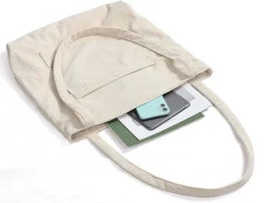 A cream-colored corduroy tote bag, seen from above with its top open, revealing a mint green smartphone and a white tablet or notebook inside. The tote has a soft, ribbed texture and a simple design, with one exterior slip pocket visible. It showcases the practical use of a tote bag for carrying daily essentials.