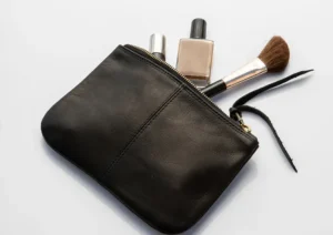 A small black leather cosmetic pouch, a popular type of makeup bag, is shown open with a makeup brush, a bottle of foundation, and a silver cosmetic tube sticking out. The background is white.