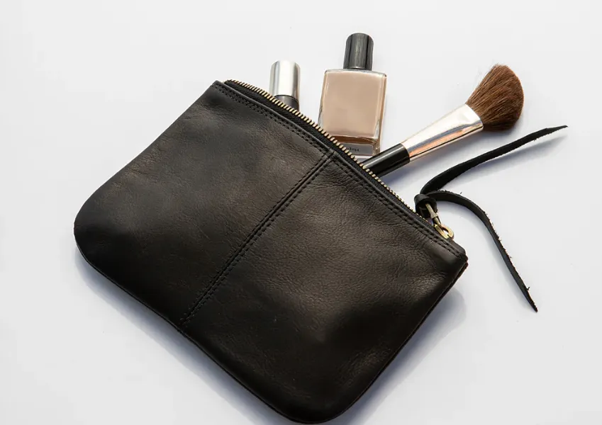 A small black leather cosmetic pouch, a popular type of makeup bag, is shown open with a makeup brush, a bottle of foundation, and a silver cosmetic tube sticking out. The background is white.