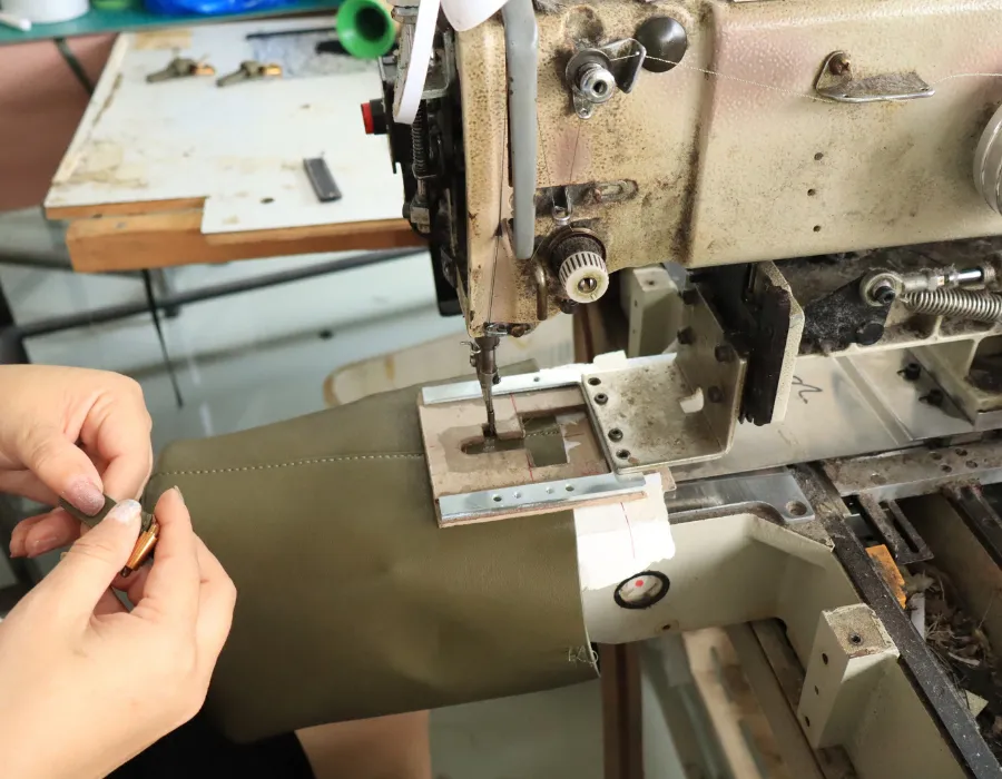 Close-up of a factory worker guiding an olive-green bag panel beneath an industrial sewing machine, with stitching visible along the edge during shoulder bag manufacturing.