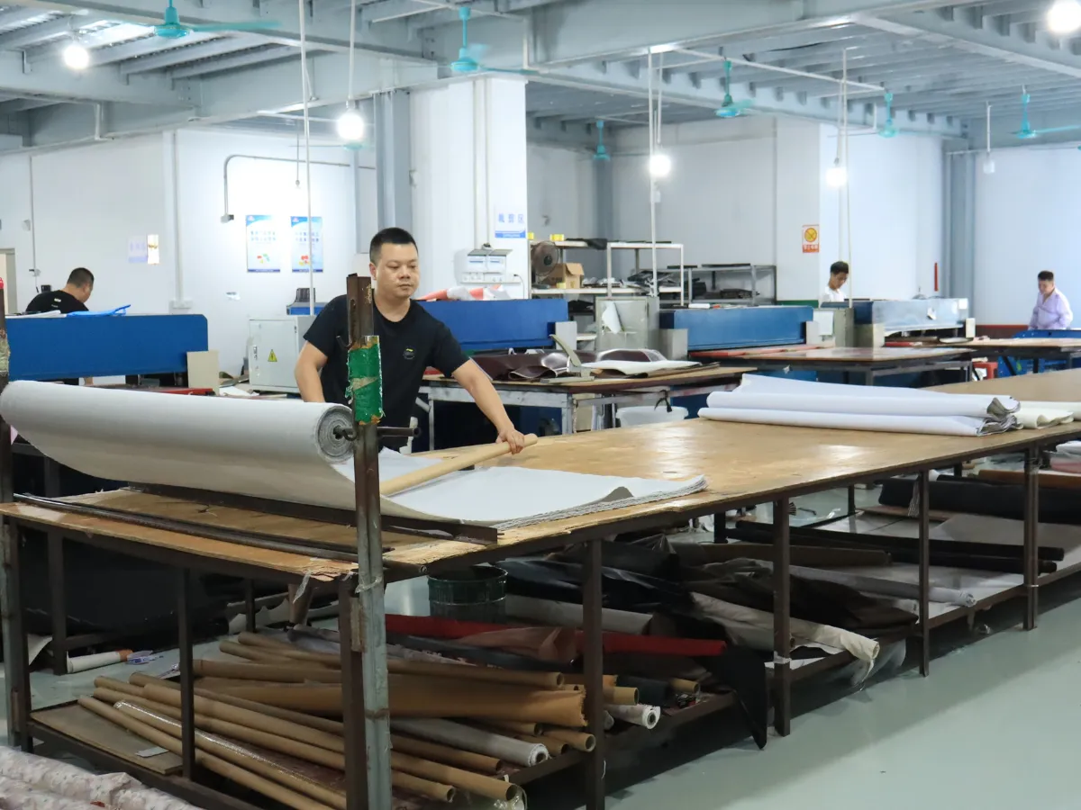 Worker preparing light-colored sheet material beside a large roll on a wooden worktable in a handbag workshop, with additional material rolls stored underneath.