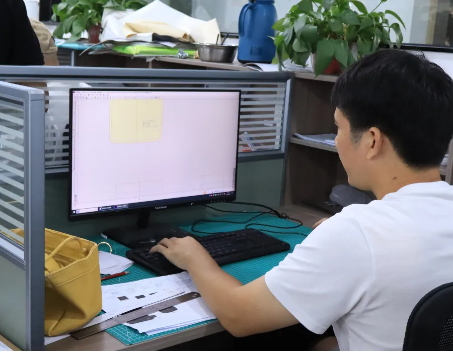 Designer working on a bag pattern displayed on a computer monitor, with a yellow bag sample and reference sheets on the desk during OEM shoulder bag construction planning.