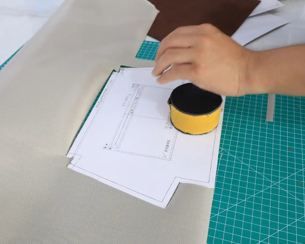 Close-up of a worker positioning a paper bag pattern on light-coloured material beside a green cutting mat, with a black and yellow pattern weight holding it in place during private label shoulder bag construction planning.