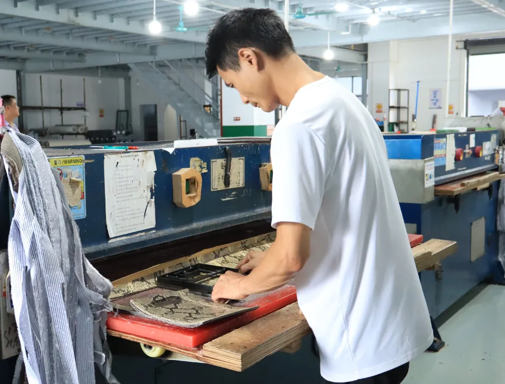 Factory worker positioning cutting dies on the work bed of an industrial cutting press to prepare bag panels and components for OEM shoulder bag production.
