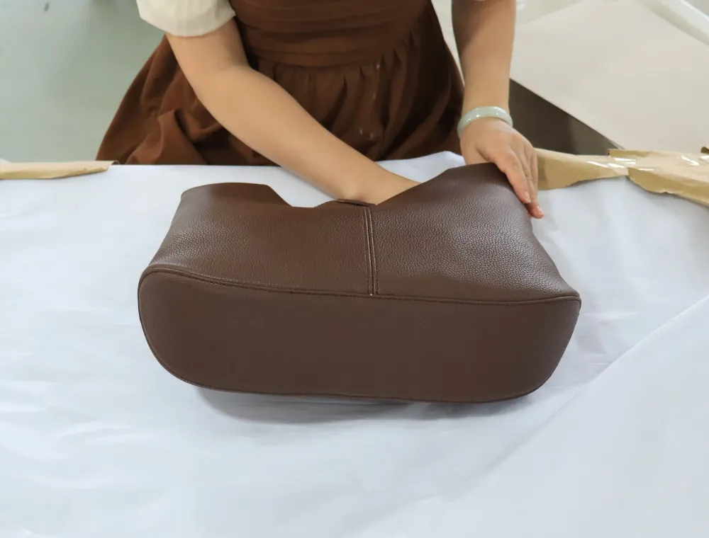 Worker checking the shape and seams of a brown textured bag on a white-covered worktable, with its rounded base facing the camera during shoulder bag finishing and quality inspection.