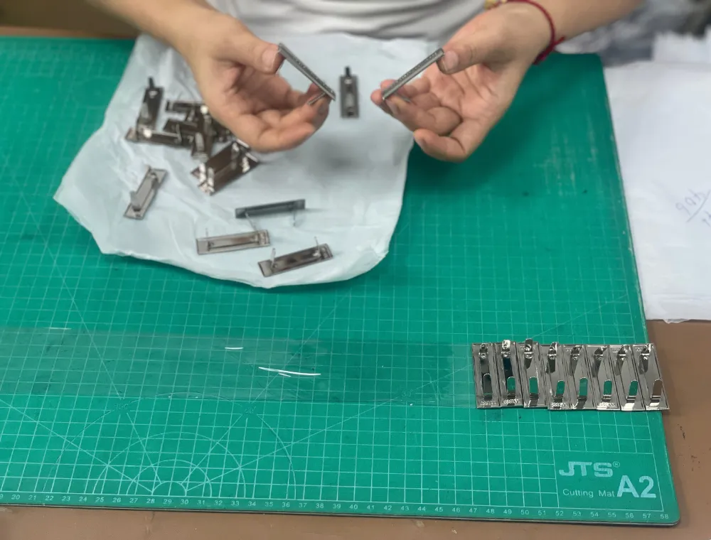 Close-up of a worker holding two silver-tone rectangular bag hardware pieces above a white cloth on a green cutting mat, with matching components nearby for private label shoulder bag hardware assembly.