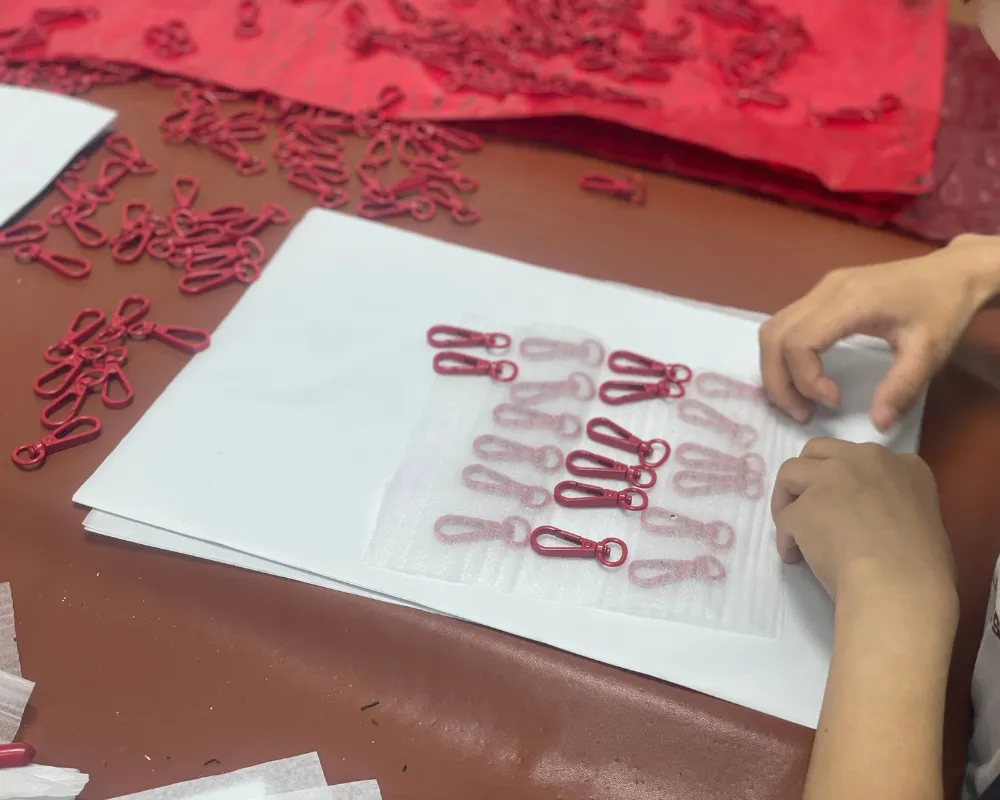 Worker arranging red bag hardware components in rows on white sheets, with additional matching pieces on the worktable during OEM shoulder bag hardware preparation.
