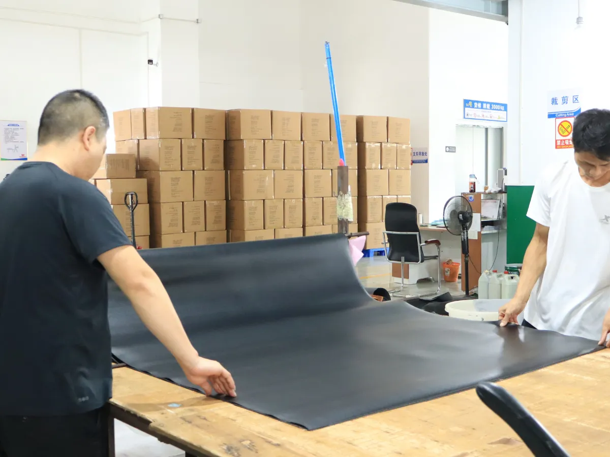 Two factory workers spreading a large black material sheet across a wooden worktable, with stacked cartons and workshop equipment in the background.