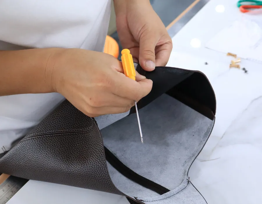 Close-up of a sample maker using a yellow-handled pointed tool on a black textured bag panel with its grey underside exposed during private label shoulder bag sample assembly.