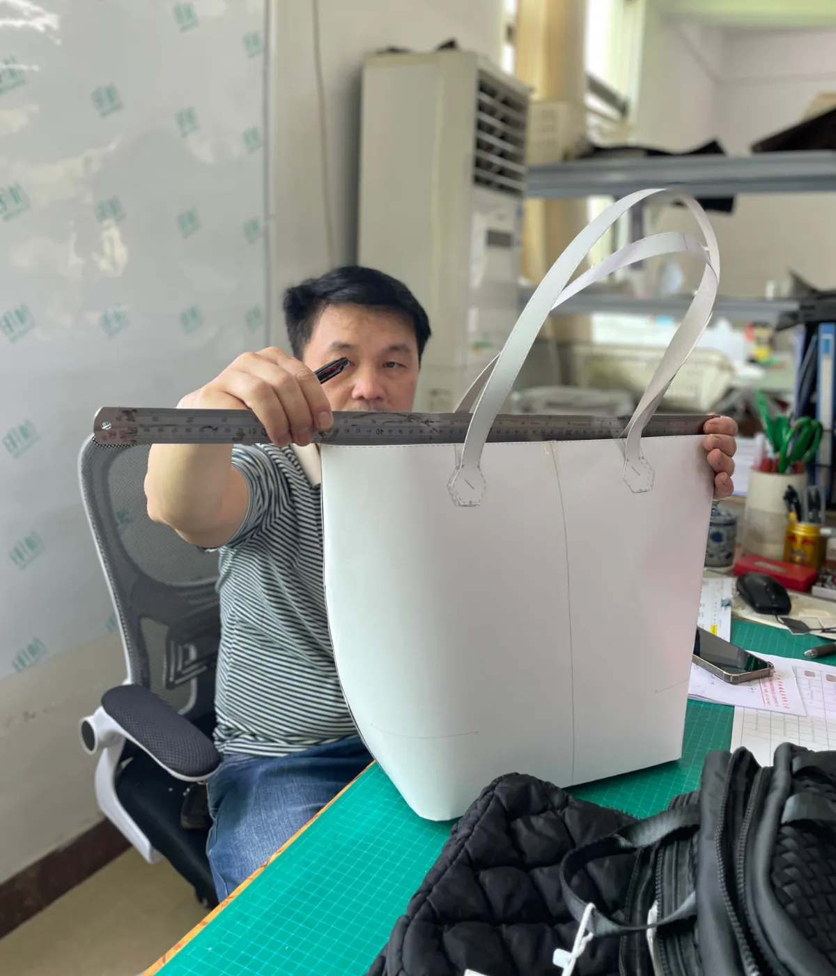 Sample maker measuring the top width of a white bag prototype with long handles using a metal ruler on a green cutting mat during custom shoulder bag development.