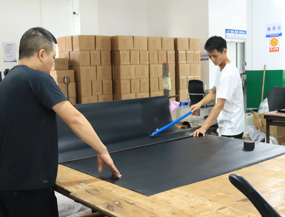Two factory workers spreading and checking a large black material sheet on a wooden table, with stacked cartons behind them during custom shoulder bag material inspection.