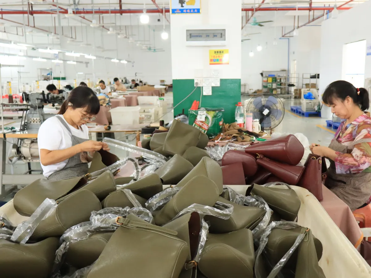 Factory workers handling olive-green and burgundy handbags at a production table piled with bags, with protective wrapping around many of the handles.