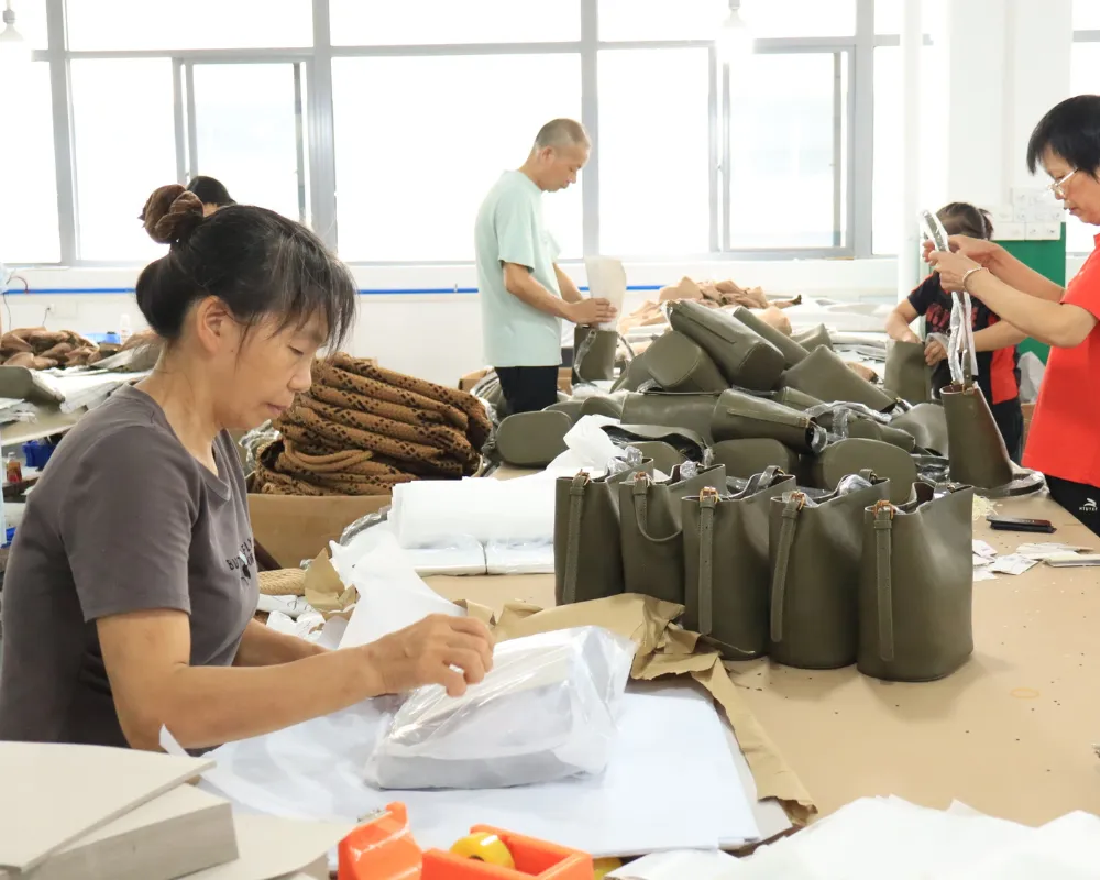 Factory workers handling olive-green handbags at a finishing and packing table, with completed bags arranged in rows and packing materials nearby for wholesale shoulder bag order preparation.