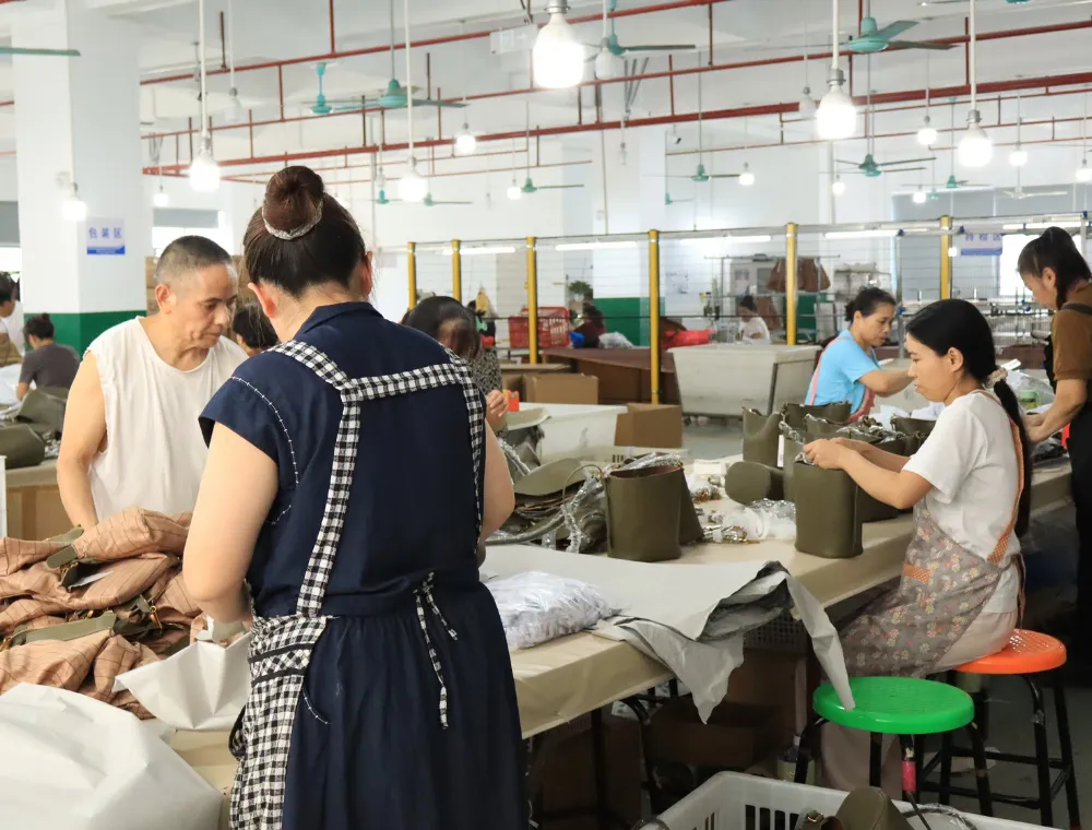 Factory team handling brown and olive-green bags at a finishing and packing worktable, with white protective materials and cartons nearby during bulk shoulder bag order preparation.