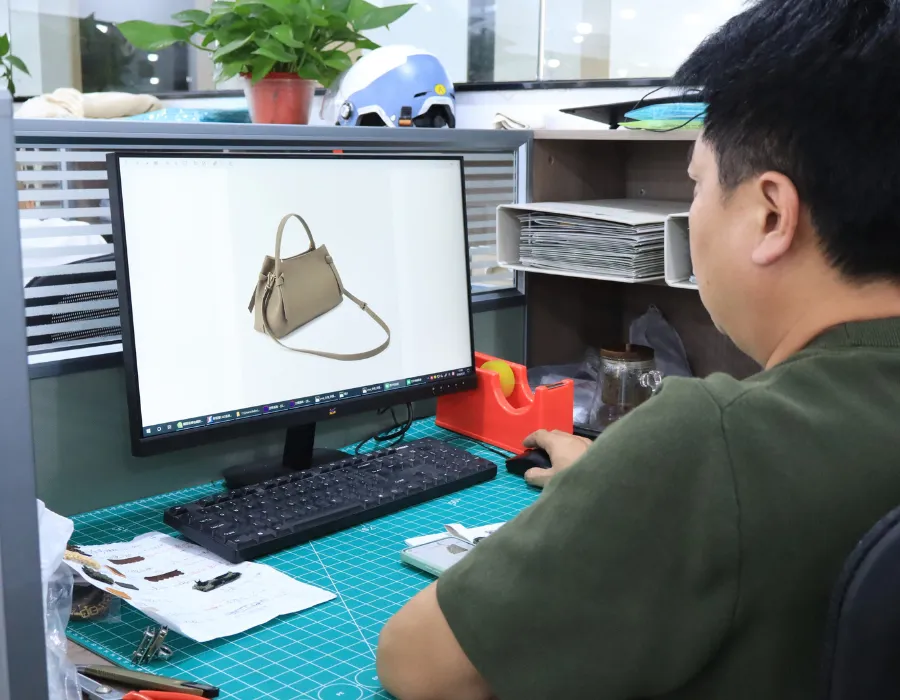 Designer reviewing a beige handbag reference with short handles and a long strap on a computer monitor at a worktable with a green cutting mat during custom shoulder bag brief review.