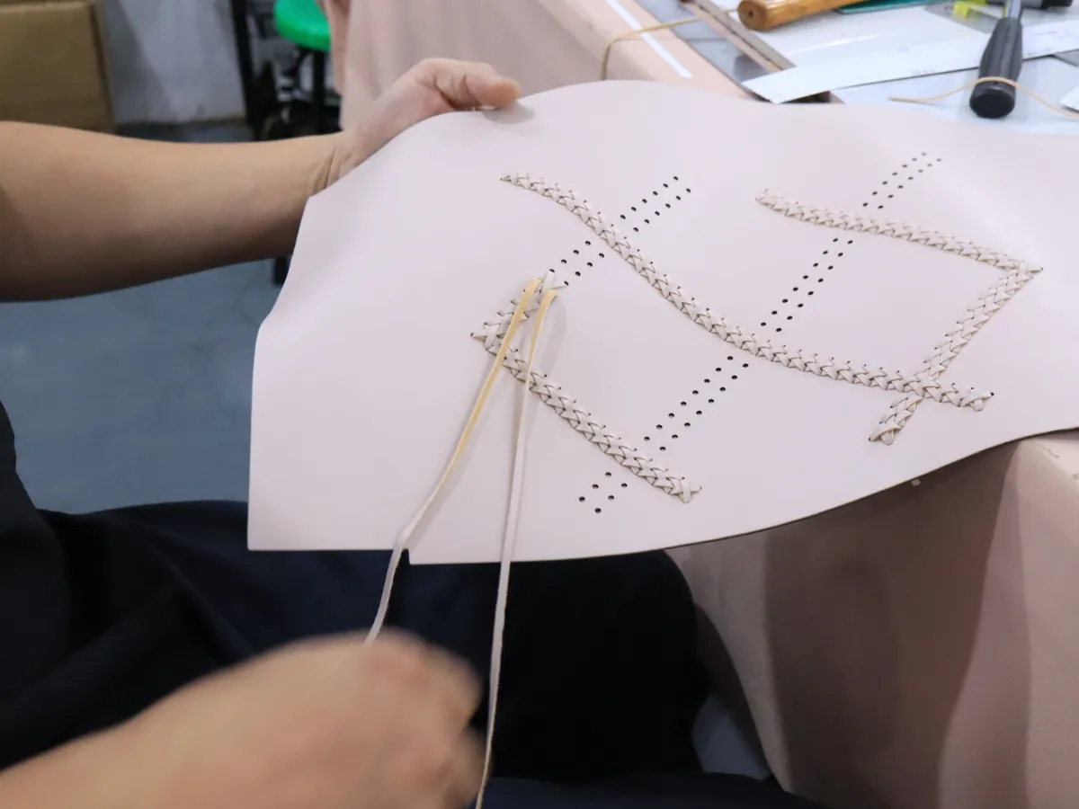 Close-up of a worker threading narrow beige strips through rows of punched holes in a pale pink bag panel to create decorative interlaced patterns.